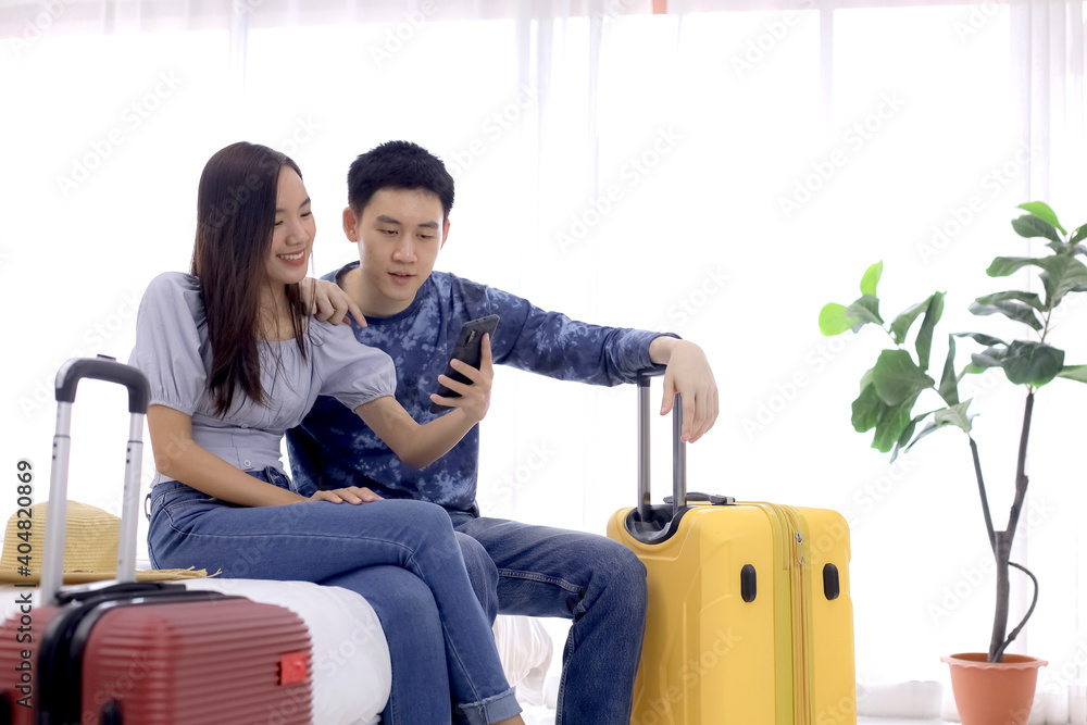 Young couple using mobile phone while sitting together on bed at a hotel room with a suitcase. Couple of tourists searching location by smartphone on bed at resort. Honeymoon trip scene. Accommodation Young couple using mobile phone while sitting together on bed at a hotel room with a suitcase. Couple of tourists searching location by smartphone on bed at resort. Honeymoon trip scene. Accommodation