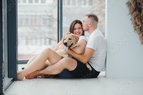 The young happy couple is moving into a new house. They are sitting down on the floor with their little puppy after they brought boxes with things to their new home.