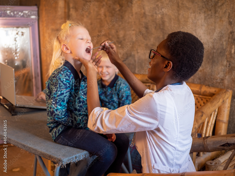 Φωτογραφία African American woman wearing white coat dentist examines blonde boy in village clinic