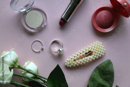 Silver rings, pearl barrette, various make up products and white roses on pale pink background. Flat lay.