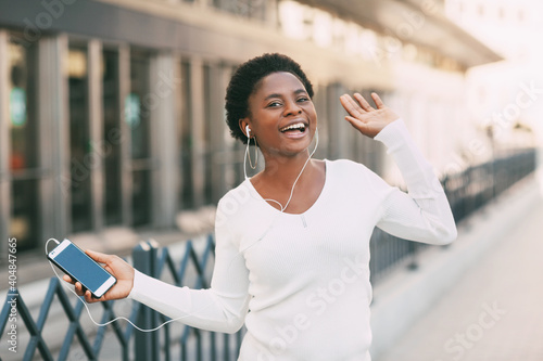 A young African woman listens to music with headphones and dances to the beat outdoors on a city street