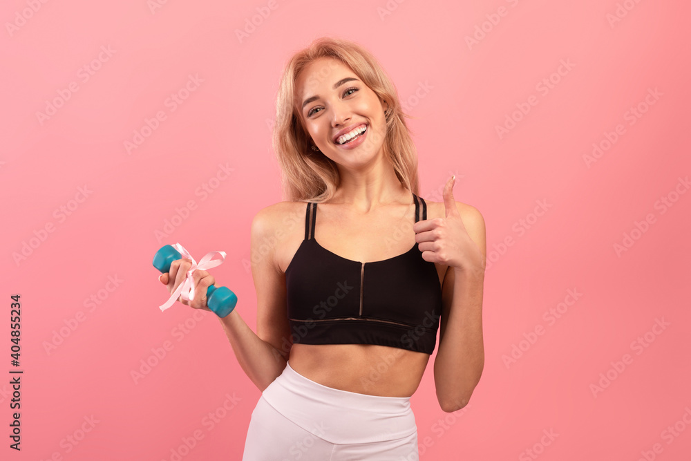 Best gift for active millennials. Fit blonde woman holding dumbbell with ribbon and showing thumb up on pink background Best gift for active millennials. Fit blonde woman holding dumbbell with ribbon and showing thumb up on pink background