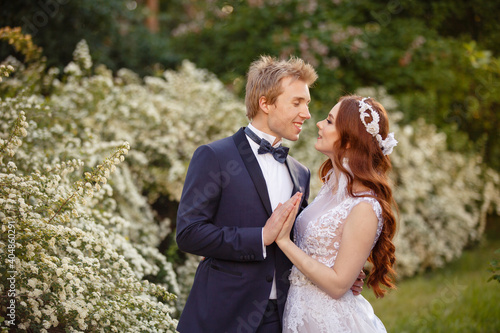 Young Bride and Groom couple in a blooming garden. Tender holding each other. Spring wedding. Redhead girl with long hairs. Young family outdoor image near blooming bush of spirea. Love and tenderness © Olga Mishyna