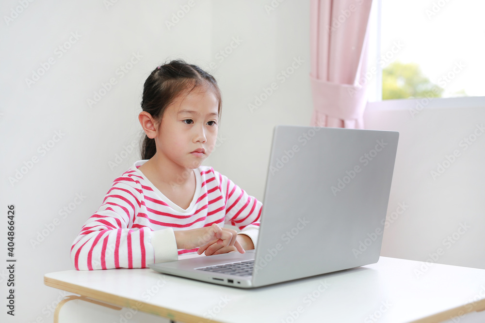 Intend asian little child sitting at desk and using laptop at home Intend asian little child sitting at desk and using laptop at home