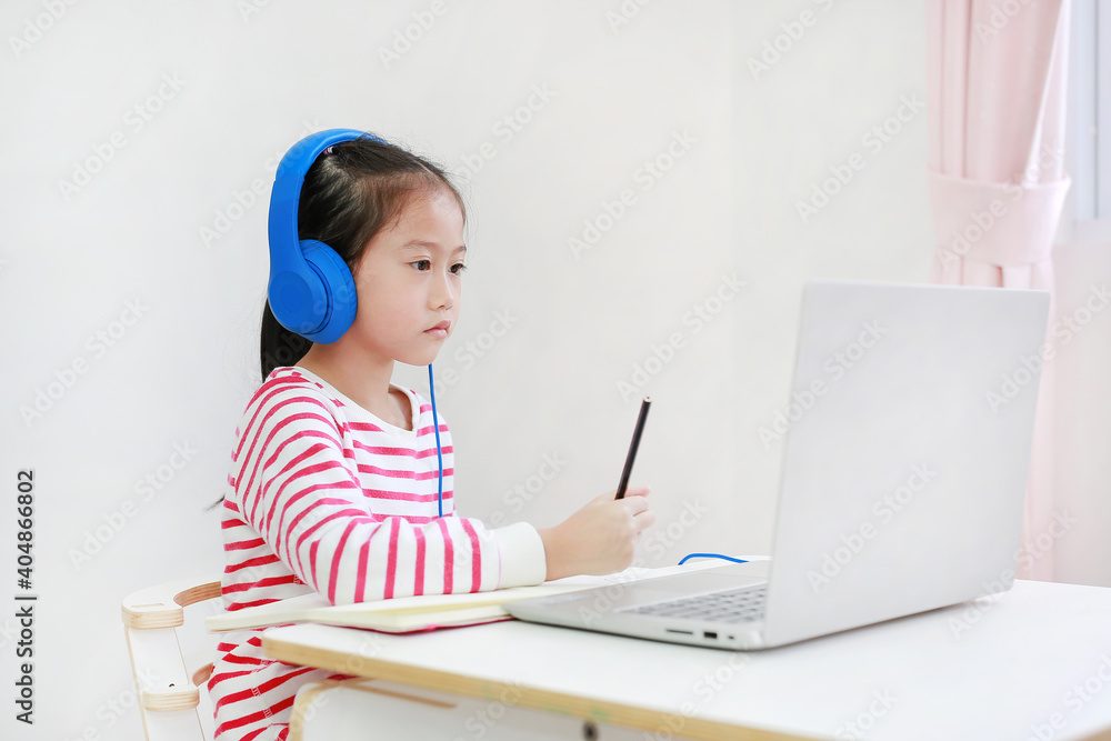 Asian little girl wearing headphone writing and study online learning class with laptop during new normal Covid-19 coronavirus at home Asian little girl wearing headphone writing and study online learning class with laptop during new normal Covid-19 coronavirus at home