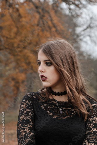 Girl in a black dress and accessories posing in a gloomy autumn garden. The brunette looked away. Black makeup in the Gothic style. Witch's photo shoot for Halloween.