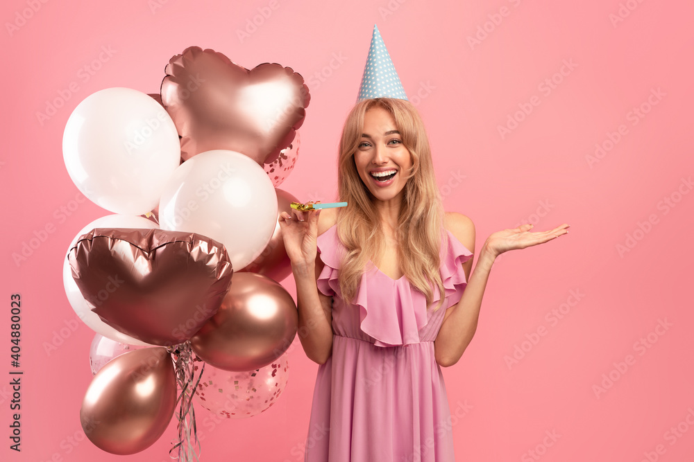 Young blonde lady with festive balloons, birthday cap and party blower celebrating special occasion on pink background Young blonde lady with festive balloons, birthday cap and party blower celebrating special occasion on pink background