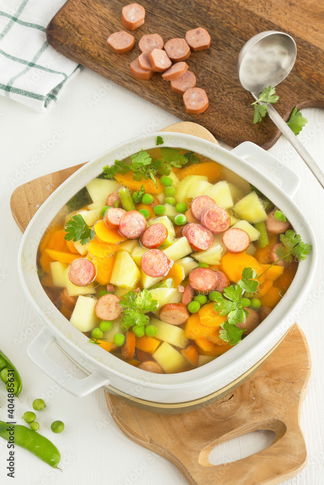 Foto Vegetable soup with sausages in a soup tureen with sausage slices on a wooden bo