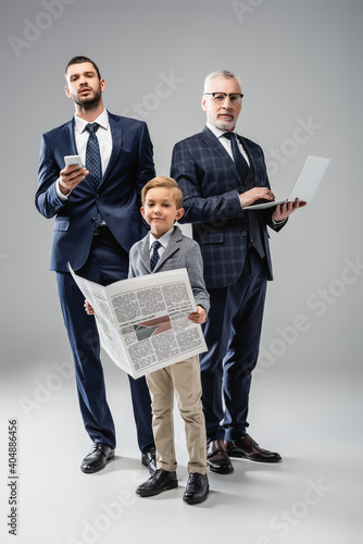 smiling boy with newspaper looking at camera near dad and grandfather holding gadgets on grey photo