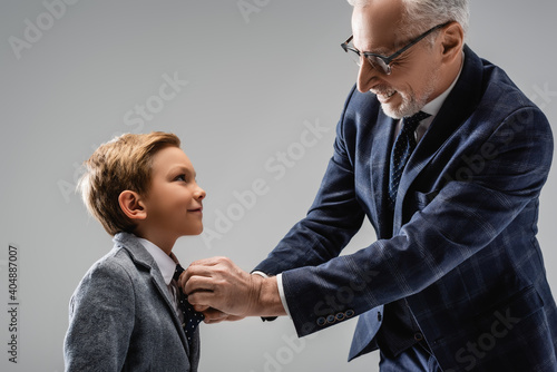 happy businessman in eyeglasses buttoning blazer of smiling grandson isolated on grey photo