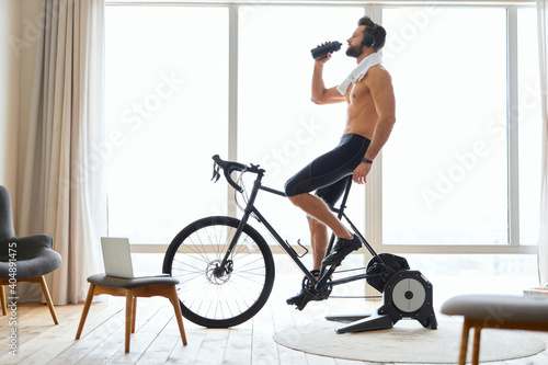 Athletic young man riding stationery bike and drinking water