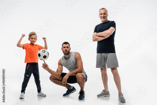 man in sportswear whistling while playing with soccer ball near boy and grandfather on white