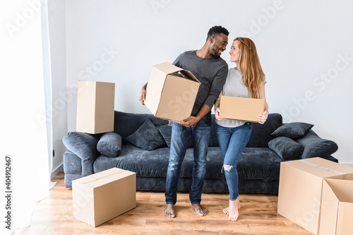 Happy newlyweds relocate to new apartment. Young married couple standing in living room, holding in arms cardboard boxes. Caucasian woman with african american man unpacking stuff, moving day concept