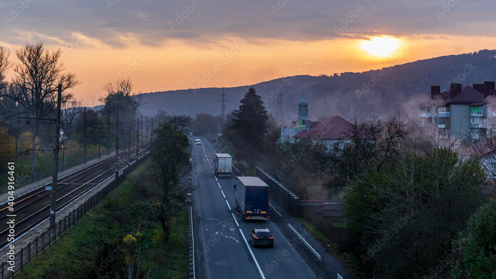 Railway track and mountain road in a small village at sunset. The sun is peeping through the storm clouds. Fog hangs in tall trees. Colorful autumn in the Ukrainian Carpathians.