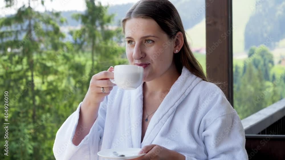 Happy smiling woan in bathrobe drinking coffee and enjoying view on mountains at hotel. Concept of people traveling, tourism and vacation in mountains at summer Happy smiling woan in bathrobe drinking coffee and enjoying view on mountains at hotel. Concept of people traveling, tourism and vacation in mountains at summer