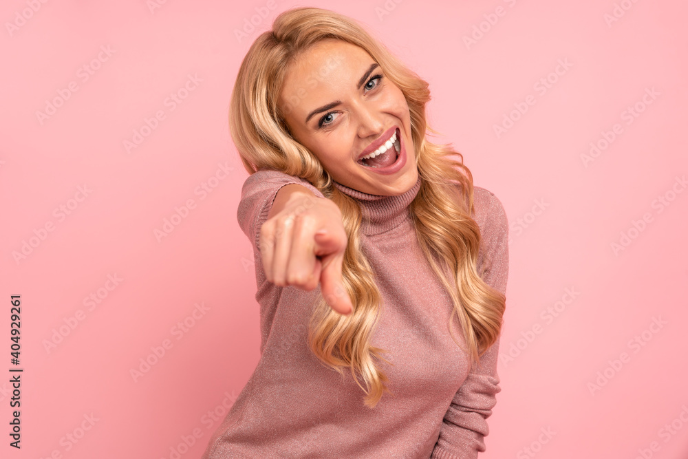 Cheerful blonde woman in sweater pointing at the camera with open mouth over pink background Cheerful blonde woman in sweater pointing at the camera with open mouth over pink background