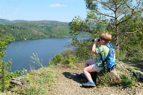 A boy looks through binoculars at a water reservoir in a valley. © Helena