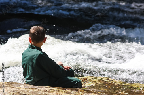 A boy in green clothes watches the flowing waters of a river sitting on a rock. Located in Batistella Reserve  Corupa  Santa Catarina State  Brazil.