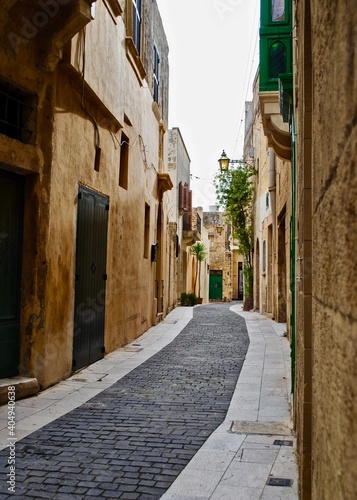 Fototapeta Naklejka Na Ścianę i Meble -  Narrow alley in Gozo, Malta with tan limestone buildings with green wood trim. 