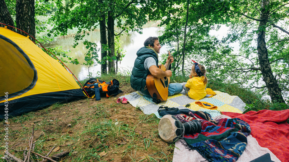 Father and his little daughter are sitting in the forest at the family camping and having fun. The father plays the guitar for his daughter. A man teaches a girl to play the guitar. Father and his little daughter are sitting in the forest at the family camping and having fun. The father plays the guitar for his daughter. A man teaches a girl to play the guitar.