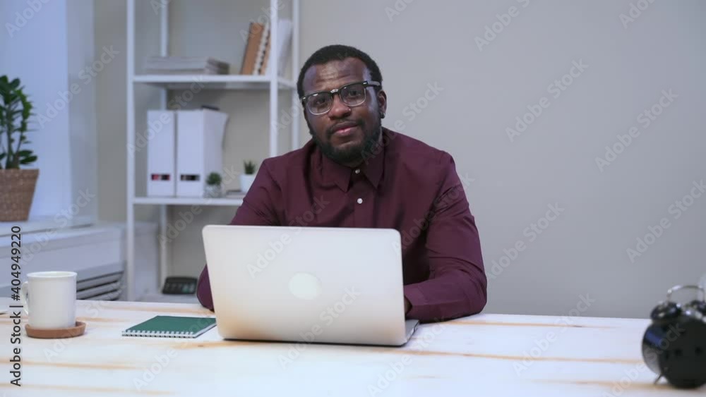 African man in glasses is working on laptop at table and smile Spbas. Black businessman with coffee and in business shirt is typing text on white computer in office. Portrait of American male model African man in glasses is working on laptop at table and smile Spbas. Black businessman with coffee and in business shirt is typing text on white computer in office. Portrait of American male model