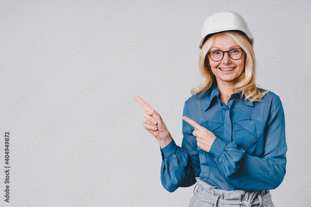 Close up portrait of a middle-aged caucasian woman pointing at copy space in hard hat and formal clothes isolated over grey background