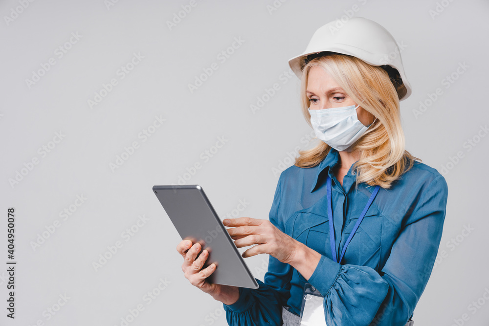 Good-looking successful aged woman using lap top in hard hat and medical mask against Covid 19 isolated over grey background Good-looking successful aged woman using lap top in hard hat and medical mask against Covid 19 isolated over grey background