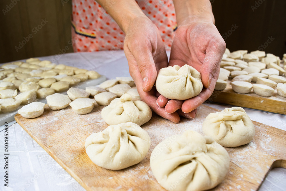 Woman's hands soiled with flour hold khinkale dumpling on the background of cutting boards with hendmade dumplings, ravioli and khinkali