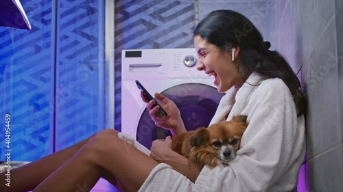 Mixed-races girl sitting on floor with Dog and watching video on smartphone while washing machines working. Multiethnic pretty women with phone resting in lundry service.