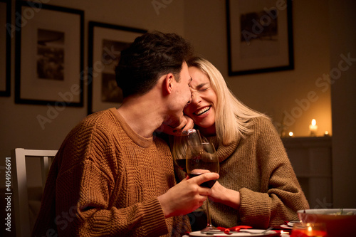 Happy young couple in love hugging, laughing, drinking wine, enjoying talking, having fun together celebrating Valentines day dining at home, having romantic dinner date with candles sitting at table. photo