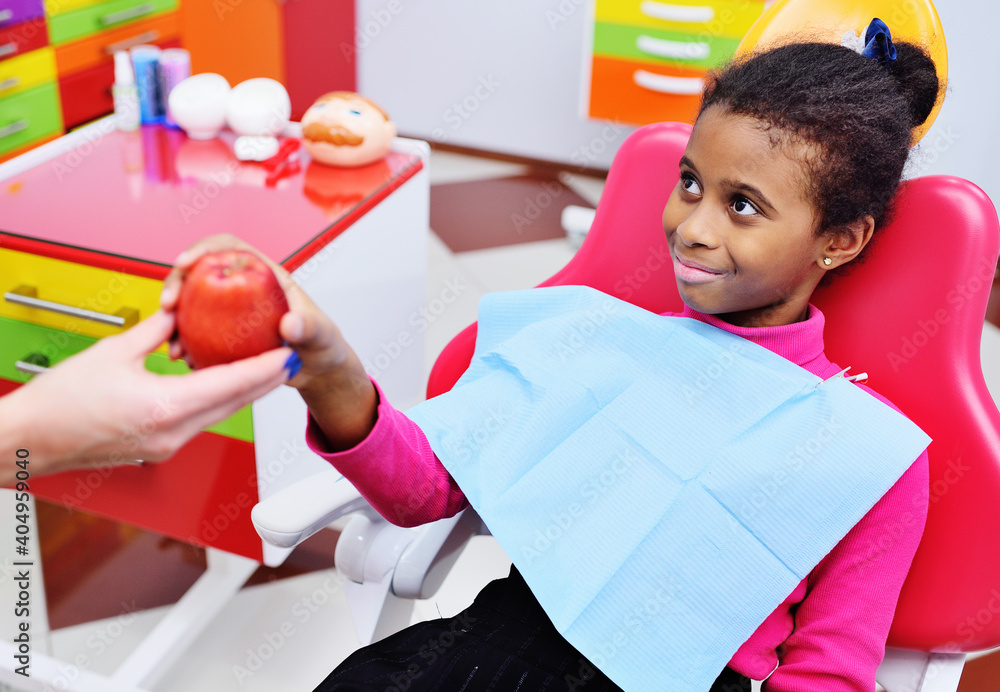 Φωτογραφία baby dentist gives a ripe red Apple to a little black African American girl in a dental chair