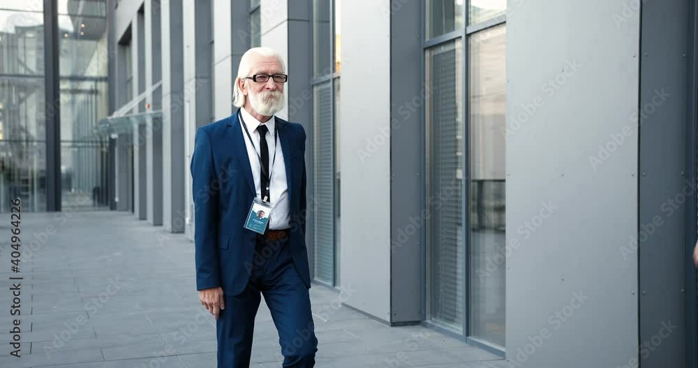 Caucasian gray-haired old man in glasses and with beard walking the street and waving with hand to male friend or colleague. Senior businessman strolling outdoors and meeting acquainted person. Caucasian gray-haired old man in glasses and with beard walking the street and waving with hand to male friend or colleague. Senior businessman strolling outdoors and meeting acquainted person.