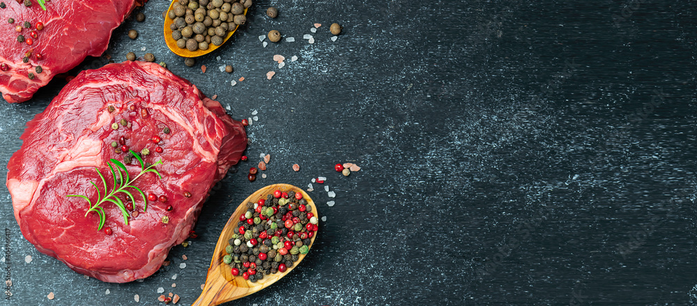 Fresh raw beef steak with assortment of spices on black rustic table long banner format.