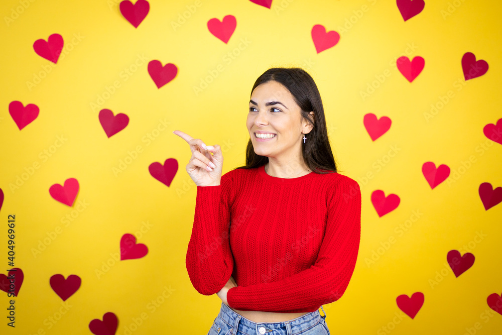 Young caucasian woman over yellow background with red hearts confused and pointing with hand and finger to the side Young caucasian woman over yellow background with red hearts confused and pointing with hand and finger to the side