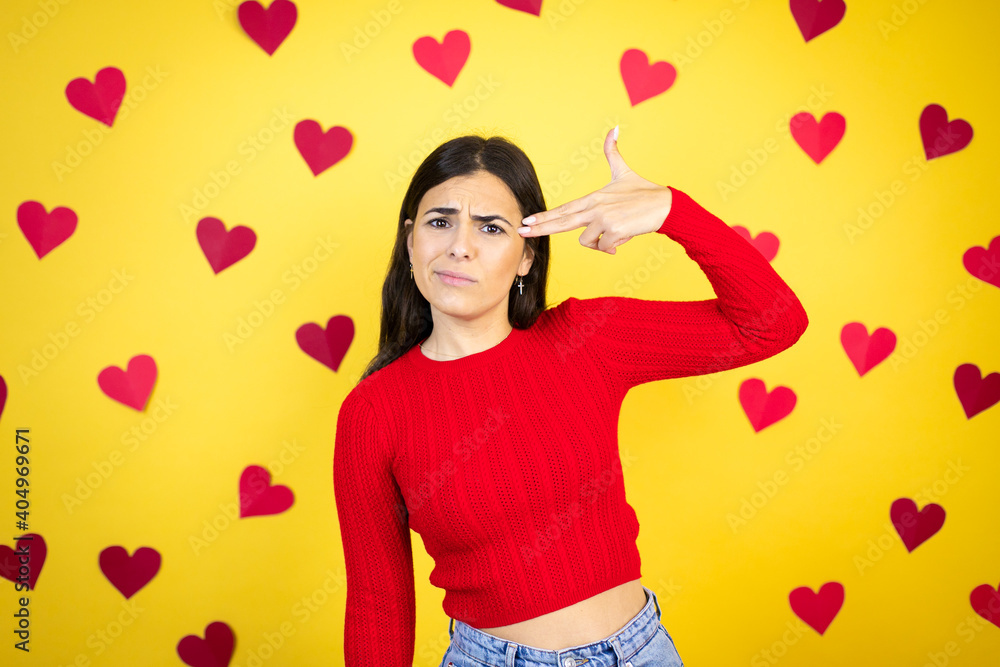 Young caucasian woman over yellow background with red hearts Shooting and killing oneself pointing hand and fingers to head like gun, suicide gesture. Young caucasian woman over yellow background with red hearts Shooting and killing oneself pointing hand and fingers to head like gun, suicide gesture.