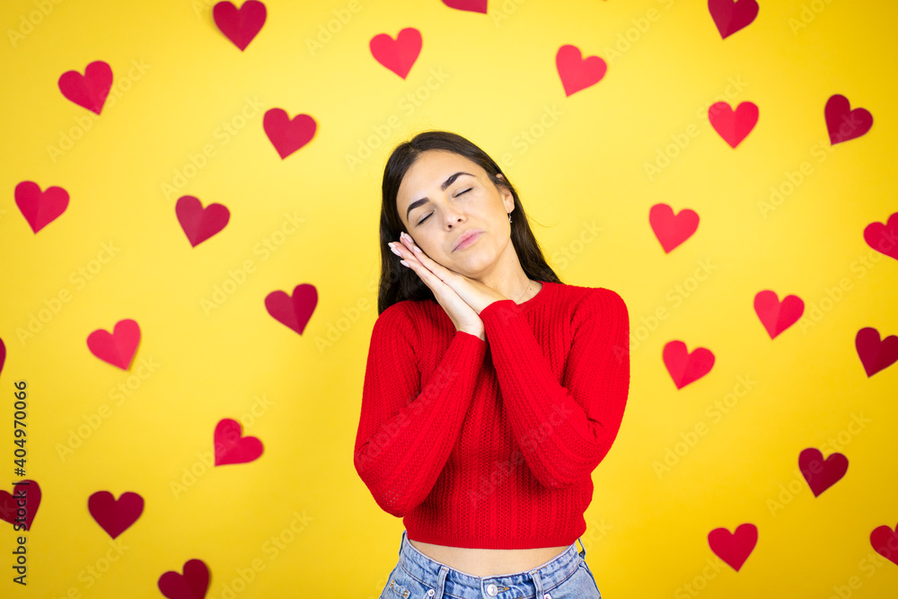 Young caucasian woman over yellow background with red hearts sleeping tired dreaming and posing with hands together while smiling with closed eyes. Young caucasian woman over yellow background with red hearts sleeping tired dreaming and posing with hands together while smiling with closed eyes.
