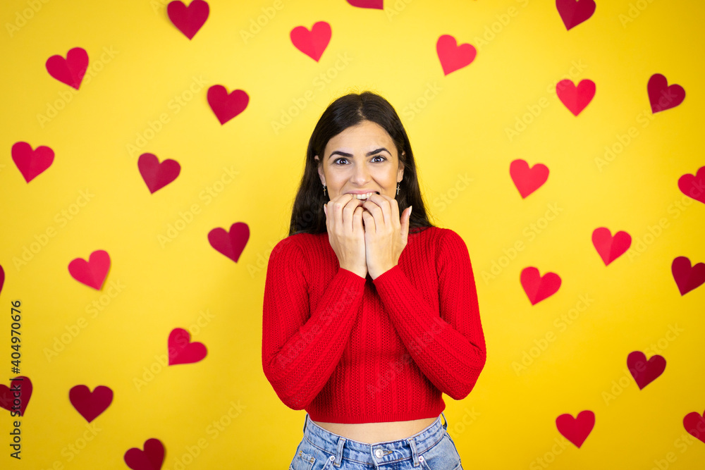Young caucasian woman over yellow background with red hearts looking stressed and nervous with hands on mouth biting nails. anxiety problem. Young caucasian woman over yellow background with red hearts looking stressed and nervous with hands on mouth biting nails. anxiety problem.