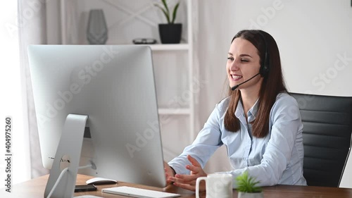 Happy young woman with a headset talking online via video connection on the trendy PC. A businesswoman with handsfree garniture waving into webcam with a pleasure smile