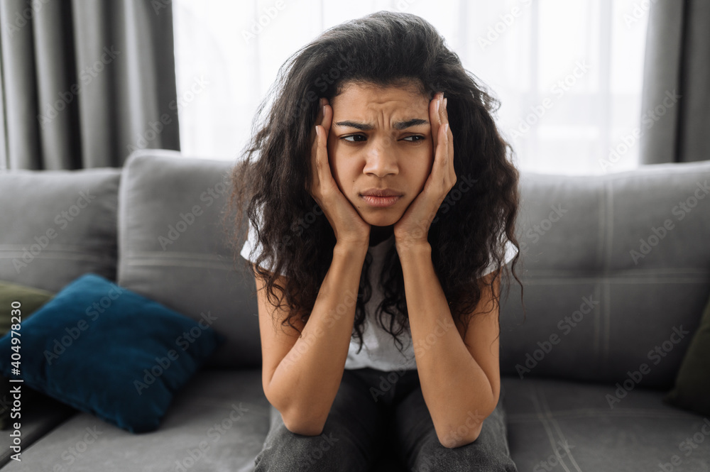 Close-up photo of a sad african american young woman. Brunette bored at home, tired or stressed. Upset girl sitting on the sofa, having a headache or feeling depressed Close-up photo of a sad african american young woman. Brunette bored at home, tired or stressed. Upset girl sitting on the sofa, having a headache or feeling depressed