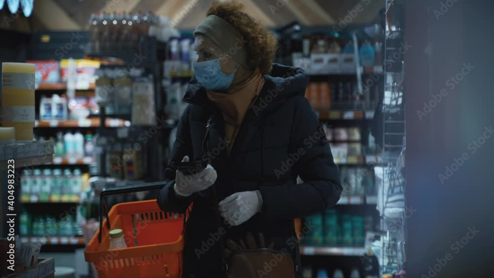 A woman shopper in a medical mask walks slowly along the shelves with the goods in search of the right ones, checking the list on the mobile phone.Safe shopping during a pandemic and an epidemic A woman shopper in a medical mask walks slowly along the shelves with the goods in search of the right ones, checking the list on the mobile phone.Safe shopping during a pandemic and an epidemic