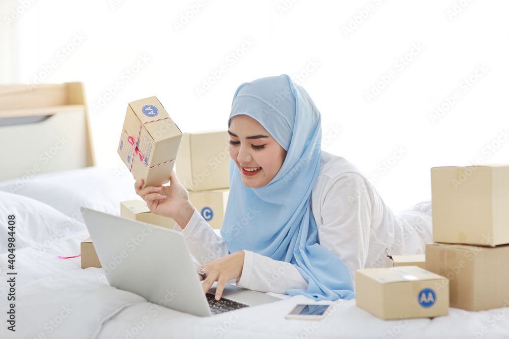 Beautiful and young asian muslim woman in sleepwear with attractive look, lies on bed with computer and online package box delivery. Startup small business SME freelance girl working with mobile phone Beautiful and young asian muslim woman in sleepwear with attractive look, lies on bed with computer and online package box delivery. Startup small business SME freelance girl working with mobile phone