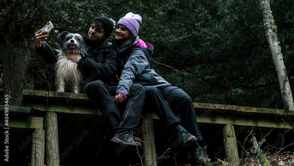 The young couple are sitting down and take out a selfie on a wooden plank road in the forest. Next to them is their shepherd dog.