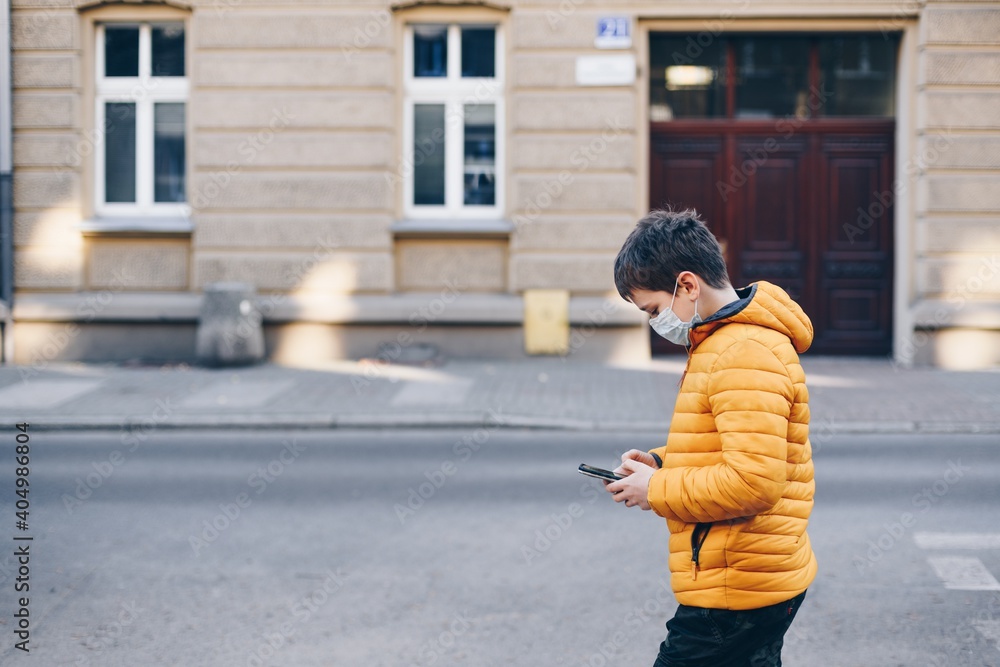 A boy in a yellow jacket wearing a protective mask is walking through the city with a phone in his hand during the pandemic.
