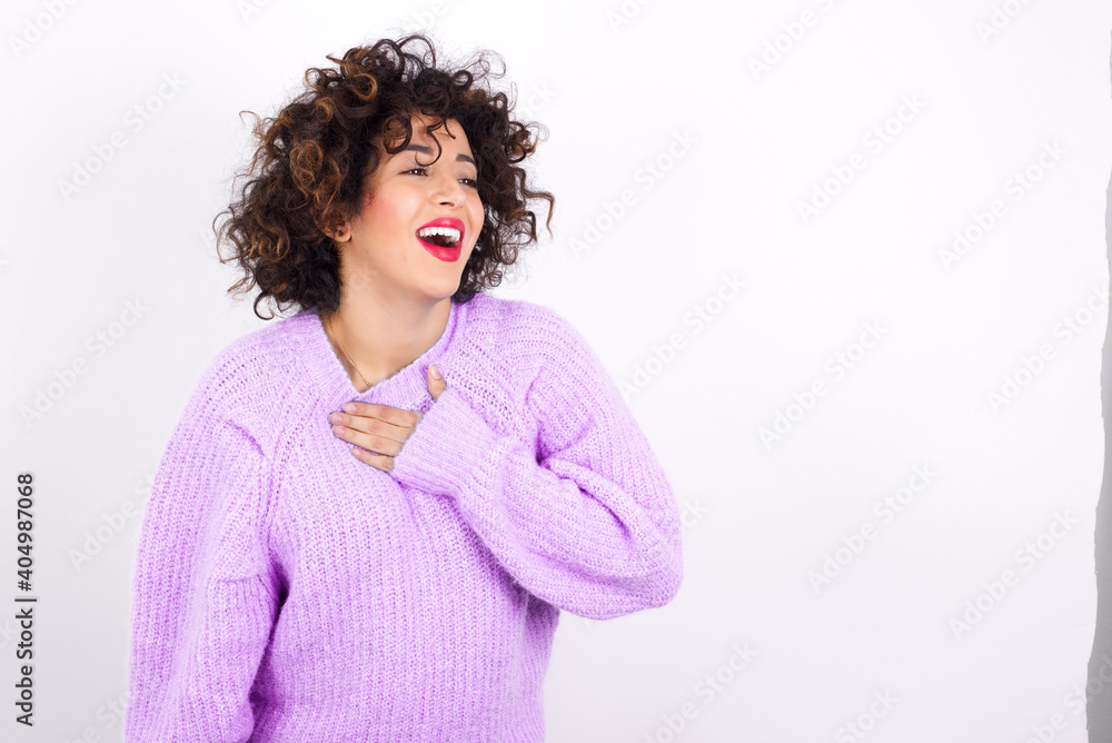 Joyful young beautiful caucasian woman wearing pink knitted sweater against white wall expresses positive emotions recalls something funny keeps hand on chest and giggles happily. Joyful young beautiful caucasian woman wearing pink knitted sweater against white wall expresses positive emotions recalls something funny keeps hand on chest and giggles happily.