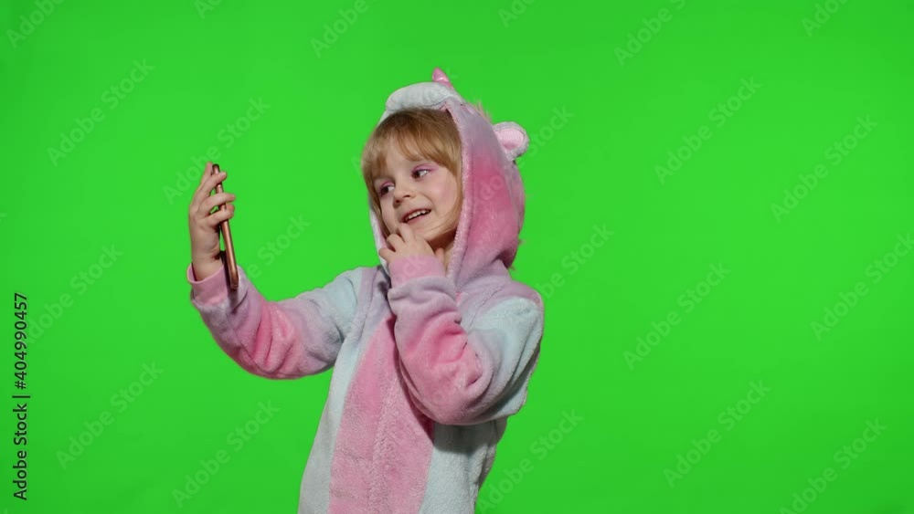 Portrait of blonde kid emotionally makes selfie on mobile phone. Child girl in unicorn pajamas costume using smartphone. Video call, blog, play games, looking at cellphone. Chroma key green background Portrait of blonde kid emotionally makes selfie on mobile phone. Child girl in unicorn pajamas costume using smartphone. Video call, blog, play games, looking at cellphone. Chroma key green background