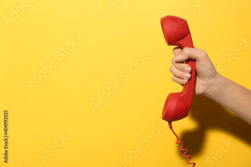 Closeup view of woman holding red corded telephone handset on yellow background, space for text. Hotline concept photo