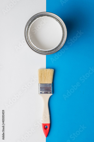 A brush and an open can of white paint on the border of white and blue colors.
