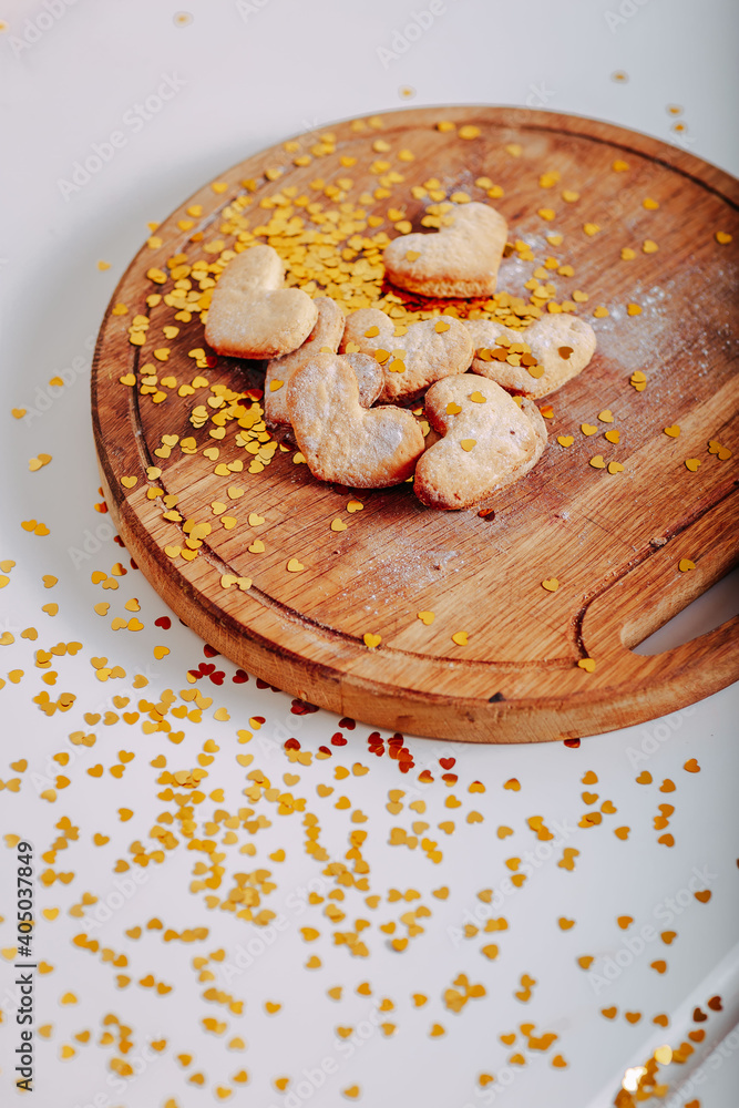 Fototapeta premium Heart-shaped cookies on round wooden cutting board. Saint Valentine's cookies in shape of heart and golden sparkles on white background. Golden glitters in shape of heart on white background.