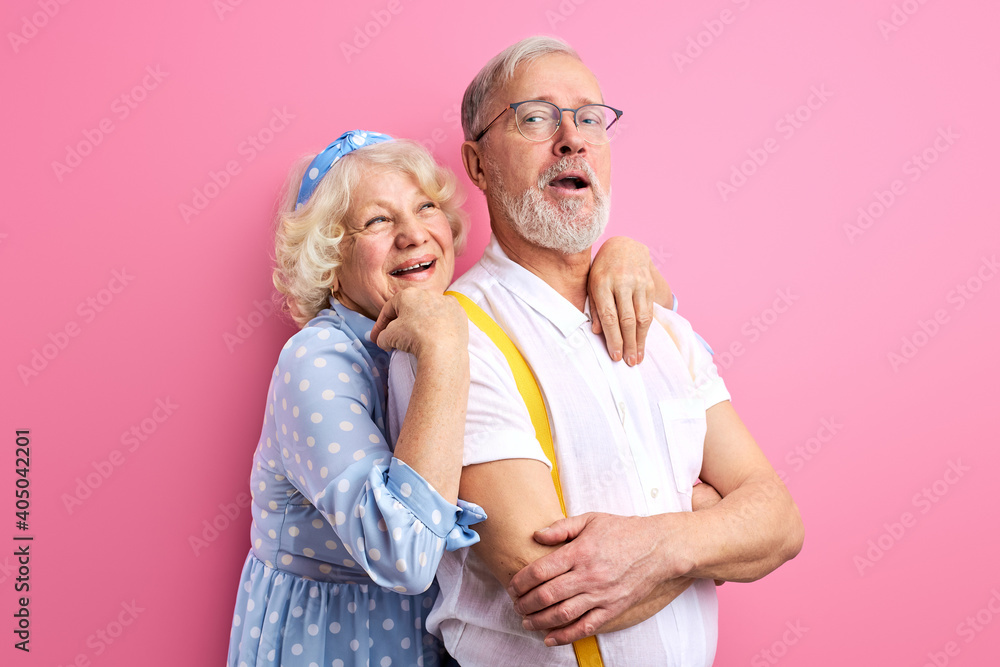 senior couple stand posing together isolated on pink background, pretty caucasian mature female hugs husband from back senior couple stand posing together isolated on pink background, pretty caucasian mature female hugs husband from back