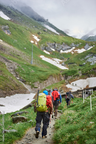 Back view of travelers with backpacks and trekking sticks walking down the road in upland valley. Group of people heading to mountain peak. Concept of traveling, hiking and mountaineering.